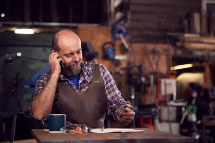 Mature Male Blacksmith Working On Design In Forge Whilst Using Mobile Phone