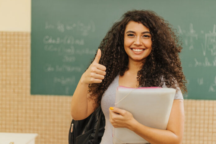 Latin Curly haired student smiling wearing backpack holding a notebook in a classroom with Thumbs up