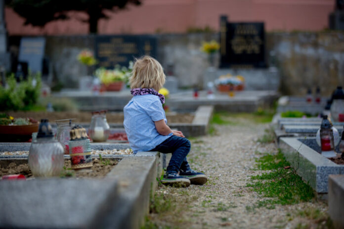 Little toddler boy, sitting on a grave in cemetery, sad and lonely, springtime